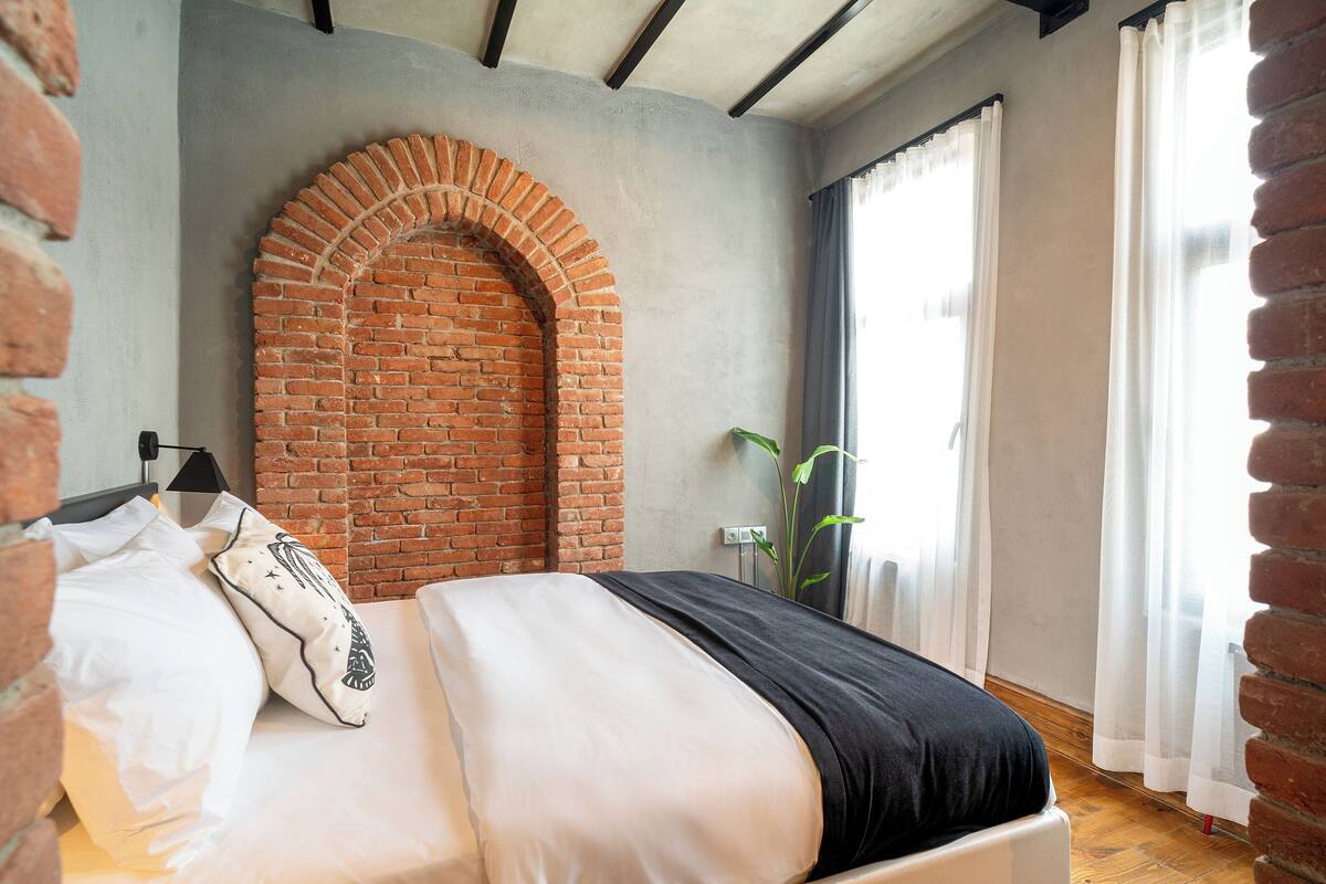 A bedroom with a brick archway, a bed with white and black bedding, a plant, and a window with sheer curtains.