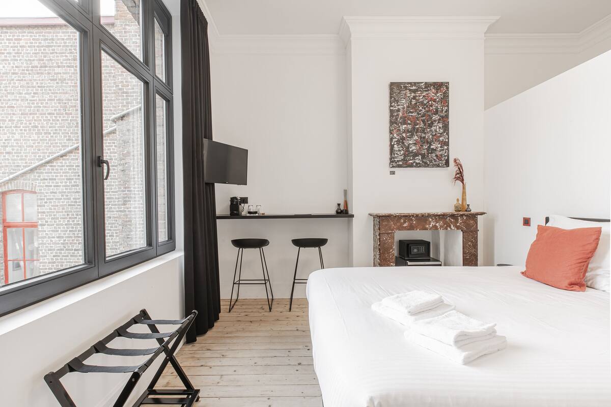 A hotel room with a bed, a desk with two stools, a fireplace, and a large window.
