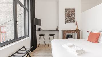 A hotel room with a bed, a desk with two stools, a fireplace, and a large window.