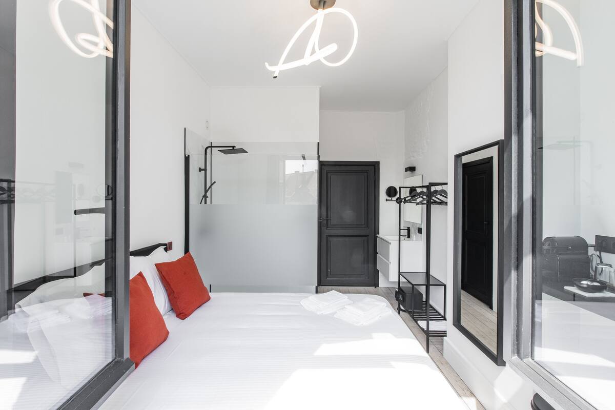A modern hotel room with a bed, red pillows, a glass shower enclosure, and a view of the outside.
