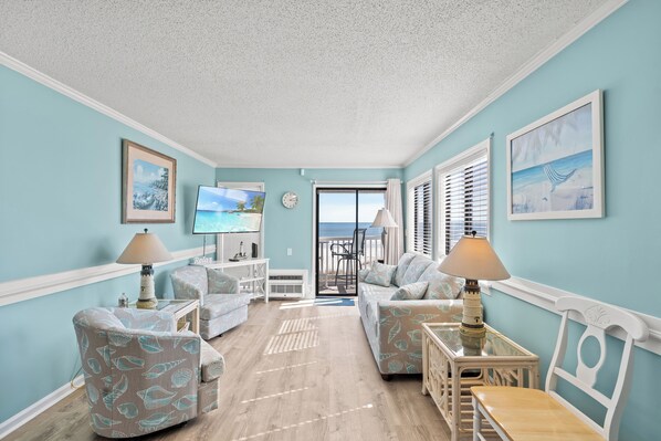 A room with light blue walls, a wooden floor, and a view to the outside. There are two sofas, a table, and a TV.