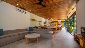 Un salon moderne avec un plafond en bois, un canapé gris, une table basse ronde et un espace cuisine visible à travers de grandes fenêtres.