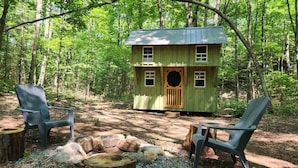 A small wooden cabin with a green roof, surrounded by trees. There are two chairs and a fire pit in front of the cabin.