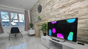 A modern living room with a large flat-screen TV, a sofa, a chair, a fireplace, and a clock on the wall. - Tyburn House, Birmingham B24 with FREE Parking and FREE WiFi (West Midlands)