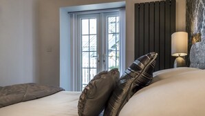 A bedroom with a bed, pillows, a nightstand with a lamp, a window with a view of buildings, and a radiator. - The Coach House - 3 Bedroom - Penally - Tenby (Tenby)