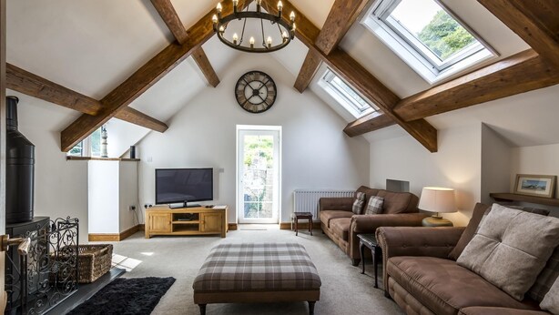 A loft-style living room with exposed wooden beams, a fireplace, a television, a sofa, and a large window. - The Coach House - 3 Bedroom - Penally - Tenby (Tenby)