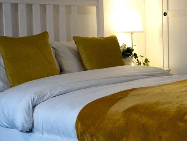 Una cama bien hecha con ropa de cama blanca y color mostaza, un cabecero blanco y una lámpara de noche.