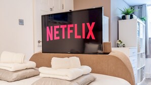 A bedroom with a bed, a TV mounted above it displaying "NETFLIX", and a white wardrobe in the background.