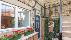 A narrow alleyway with a brick wall, a black door, and a window with red flowers.