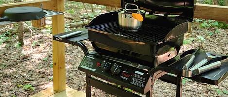 A portable grill with a gas tank and cooking utensils, set up on a wooden deck in a wooded area.