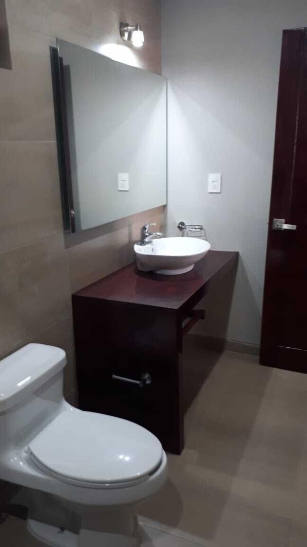 Un baño con inodoro blanco, lavamanos con espejo arriba y un mueble de madera oscura.