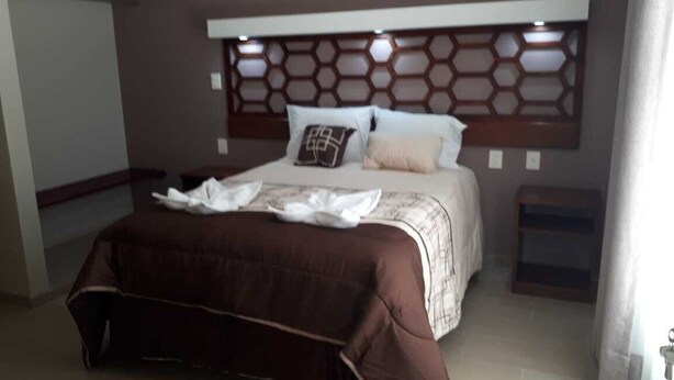 A neatly made bed with a brown bedspread and white linens, a headboard with a geometric pattern, and a bedside table with a lamp. - 9 Estrellas Hotel (Comitan de Dominguez)