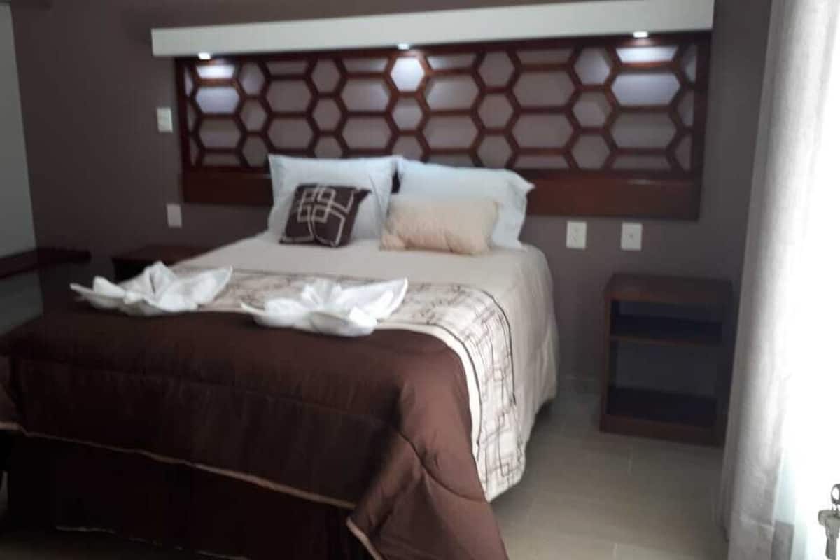 Una cama bien hecha con una colcha marrón y ropa de cama blanca, un cabecero con un diseño geométrico y una mesita de noche con una lámpara.