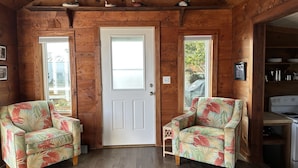 L’intérieur d’un chalet en bois, avec deux fauteuils à motifs floraux, une porte blanche et un coin cuisine.
