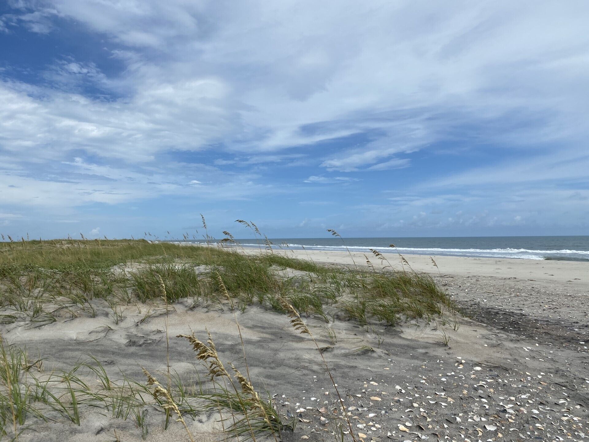A beach with sand, grass, and the ocean under a cloudy sky.