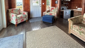 L'intérieur d'un chalet en bois, avec deux canapés à motifs floraux, un pouf bleu, un ventilateur de plafond et un coin cuisine.