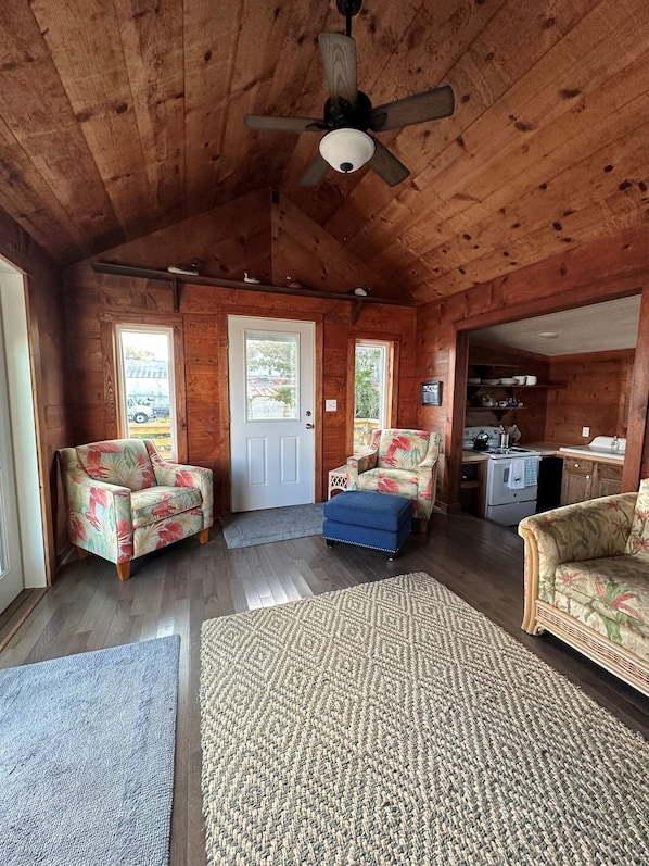 A wooden cabin interior with two floral-patterned sofas, a blue ottoman, a ceiling fan, and a kitchen area. - Charming Historic Cottage with Private Waterfront on Core Sound -Pet Friendly (Atlantic)