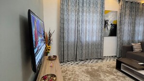 A modern living room with a flat-screen TV, a wooden coffee table, a sofa, and patterned curtains. - One bedroom apartment East legon  (Accra)