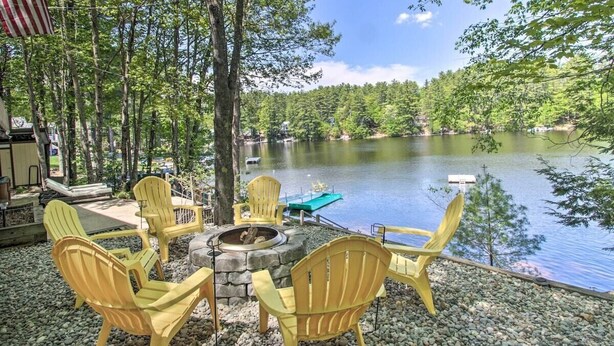 A lakeside deck with Adirondack chairs, a fire pit, and a dock extending into the water. - Stunning Lakefront Oasis—Hot Tub, Beach & Luxury! (E Wakefield)