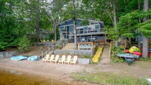 A modern house with a deck overlooking a beach, surrounded by trees. - Stunning Lakefront Oasis—Hot Tub, Beach & Luxury! (E Wakefield)