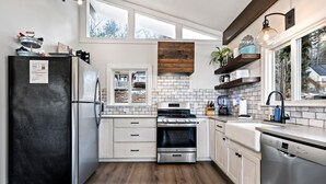 A modern kitchen with a black refrigerator, stainless steel appliances, white cabinetry, a brick backsplash, and a window overlooking the backyard. - Stunning Lakefront Oasis—Hot Tub, Beach & Luxury! (E Wakefield)