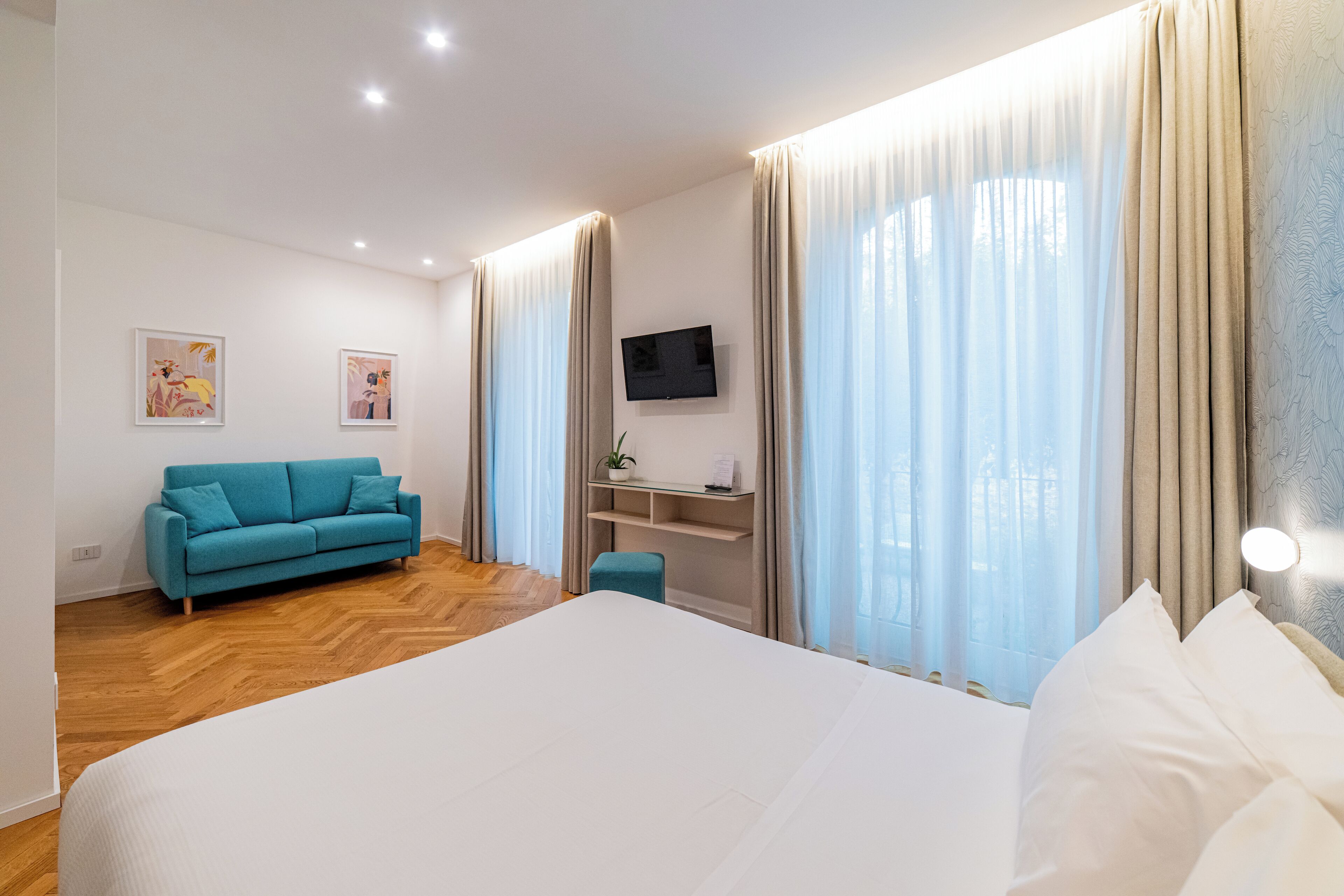 A modern hotel room with a large bed, a teal sofa, a TV mounted on the wall, and a desk with a plant.