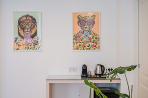Two framed artworks on a wall above a desk with a coffee maker and a plant.