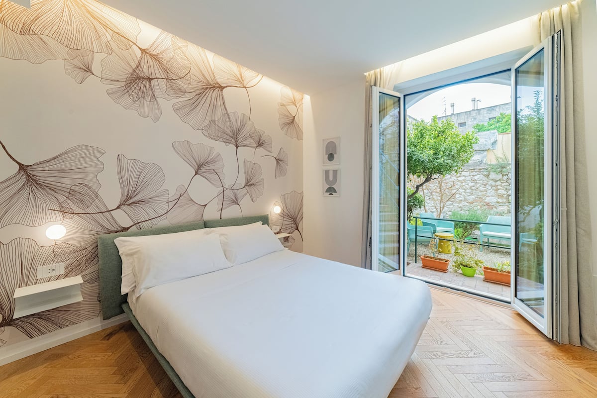 A bedroom with a bed, a nightstand, a wall with a leaf pattern, and a sliding glass door to an outdoor area with plants.