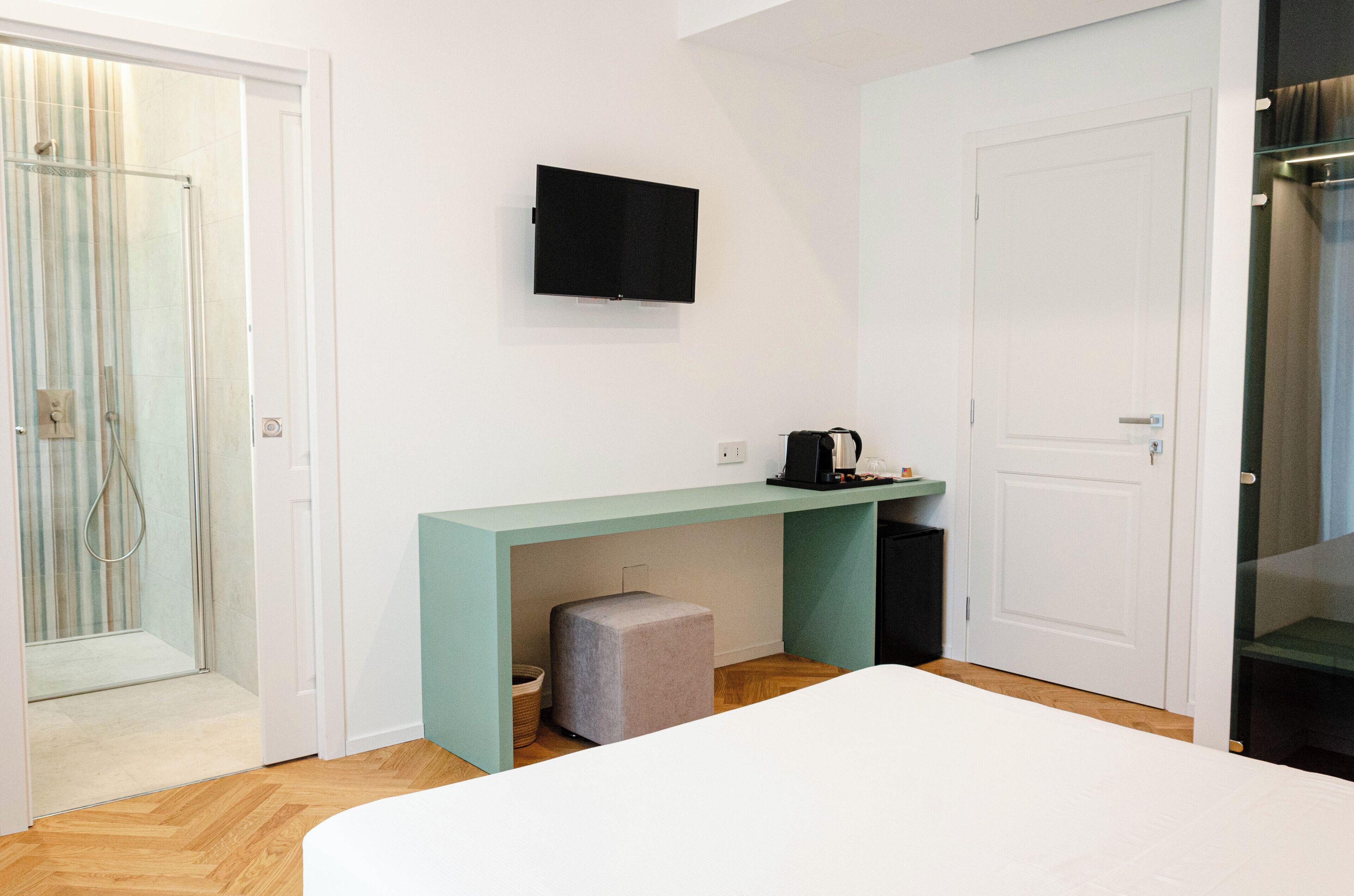 A modern hotel room with a shower, a flat-screen TV mounted on the wall, a small table with a kettle, and a bed.