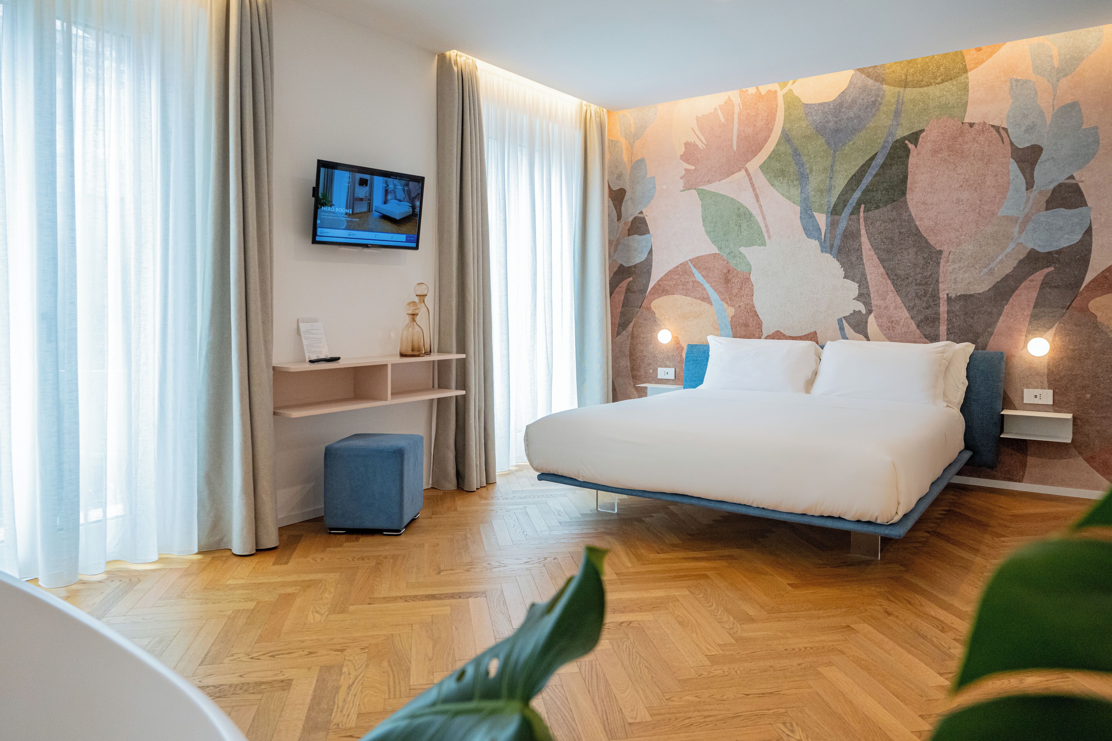 A modern hotel room with a large bed, a flat-screen TV mounted on the wall, and a floral-patterned wall.