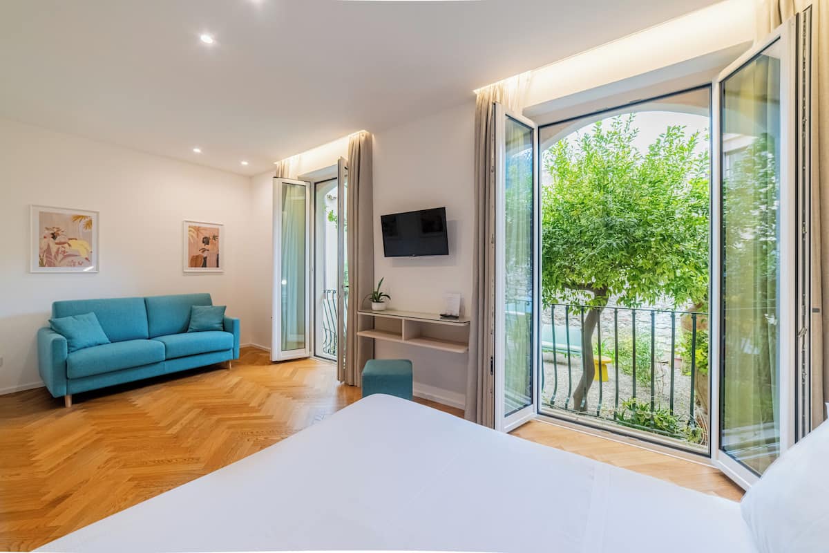A modern bedroom with a large bed, a blue sofa, a TV, and a sliding glass door to a balcony with a tree.