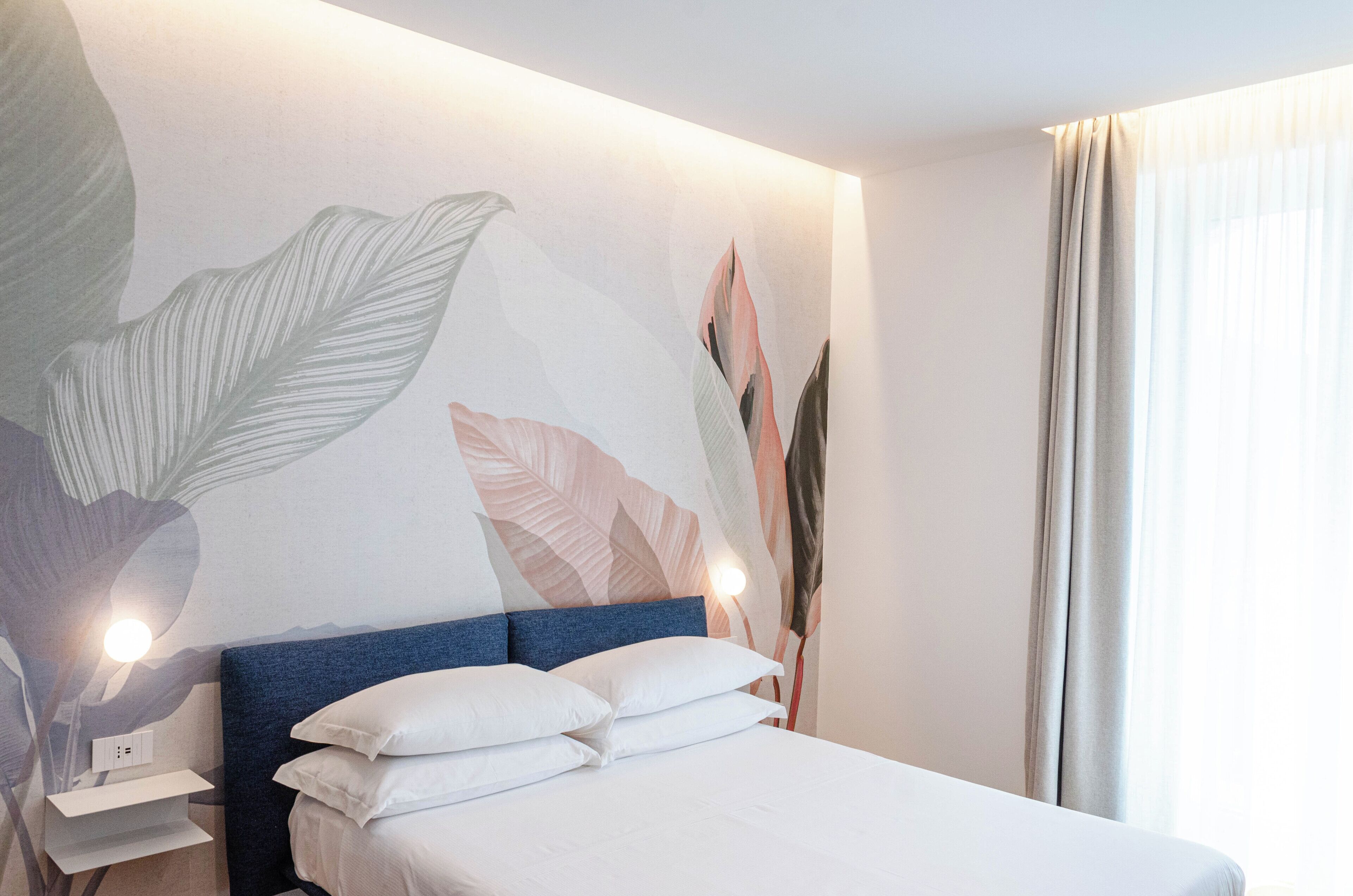 Een hotelkamer met een bed, nachtkastje, muurschildering en een raam met gordijnen.