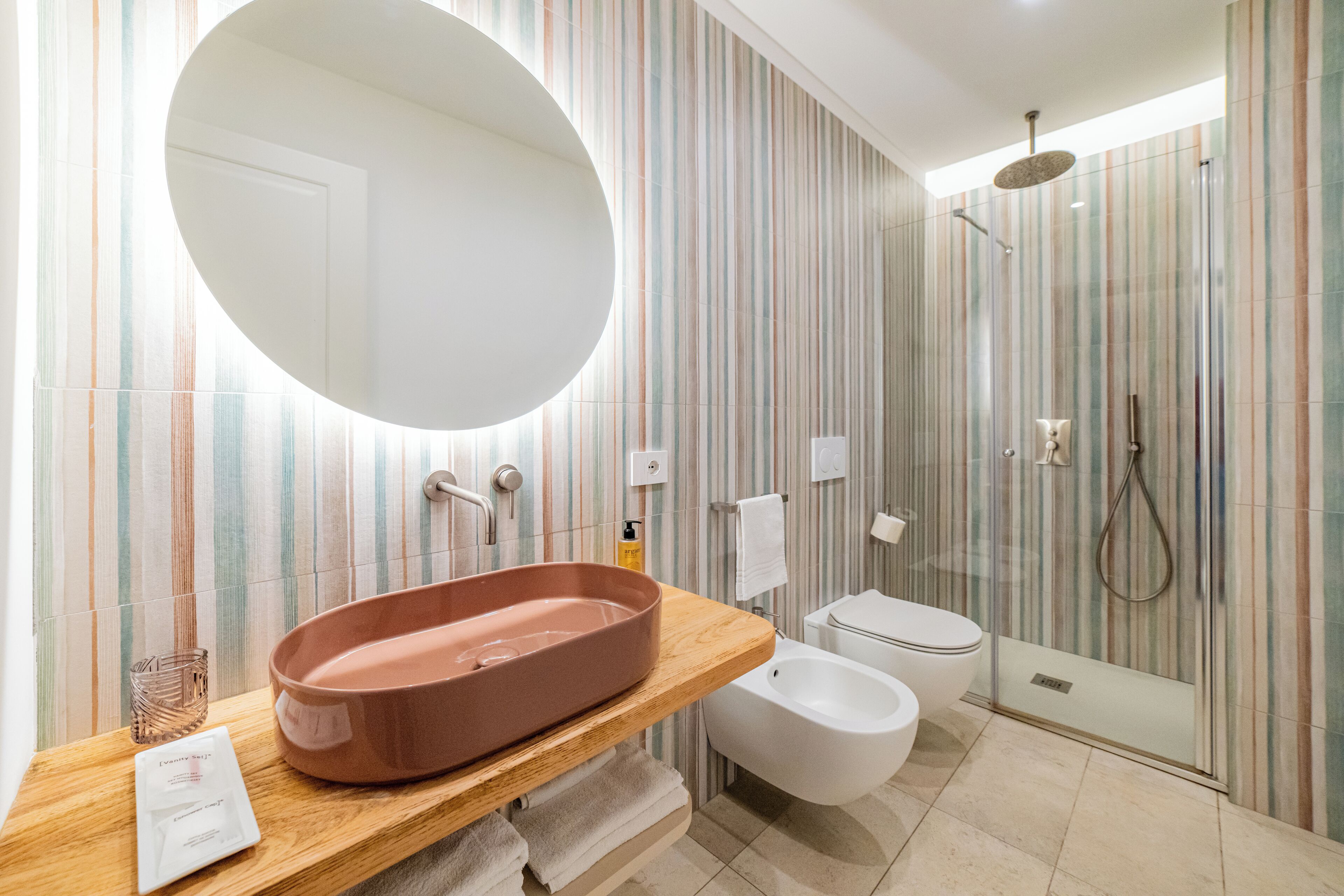 A modern bathroom with a large oval mirror, a wooden vanity, a white toilet, a bidet, and a glass-enclosed shower area.