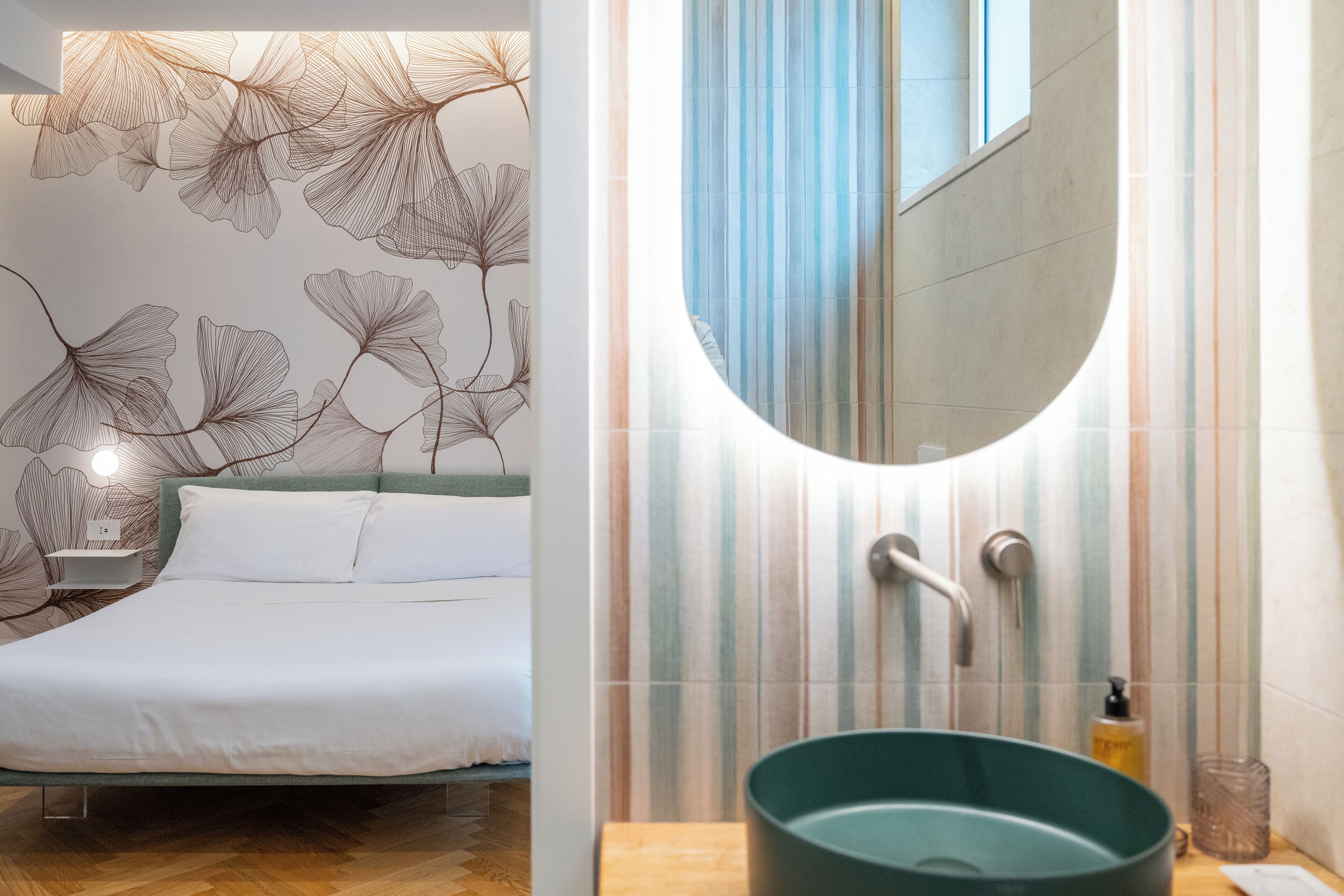 Une chambre moderne avec un lit, une table de chevet et des œuvres d’art murales. On peut également apercevoir une salle de bain avec un lavabo, un miroir et des carreaux à rayures.