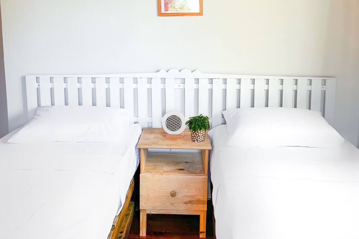 Quarto com cama branca, mesa de cabeceira de madeira, quadro na parede e um pequeno vaso com planta.