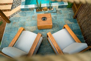 A patio with two wooden lounge chairs, a wooden table, and a pool in the background.