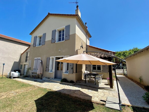 Une maison à deux étages dotée d’un patio comprenant une table et des chaises, le tout abrité sous un grand parasol.