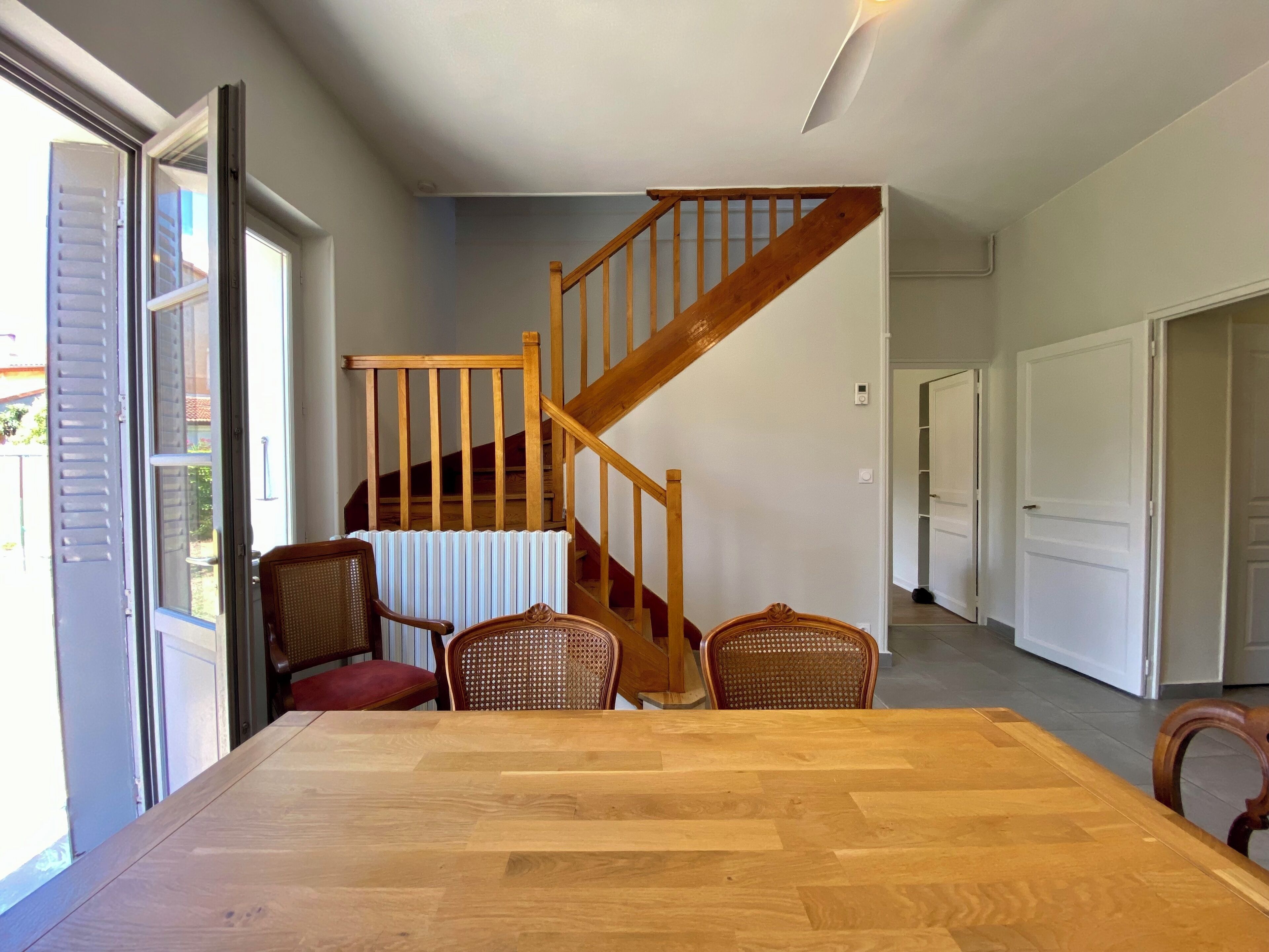 Un coin repas avec une table et des chaises en bois, un escalier et une vue sur l’extérieur par une porte ouverte.