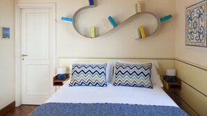 A neatly arranged hotel room with a bed, two pillows, a bedside table with a lamp, a wall-mounted shelf with books, and a decorative tile on the wall. - Resort Ravella: Lord Executive room (Sant´Agata sui Due Golfi)