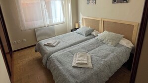 Una camera da letto con due letti, una finestra con tende leggere, un termosifone e un quadro appeso al muro.