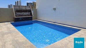 A rooftop swimming pool with a clear blue tile floor, surrounded by a low stone wall and a white building in the background. - Torre Altamar (Mazatlán)