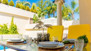 A dining table set outdoors with clear glassware, plates, and cutlery, surrounded by greenery and a pool.