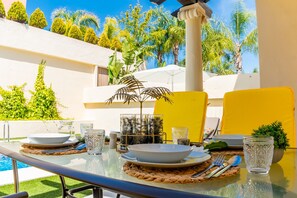 A dining table set outdoors with clear glassware, plates, and cutlery, surrounded by greenery and a pool.
