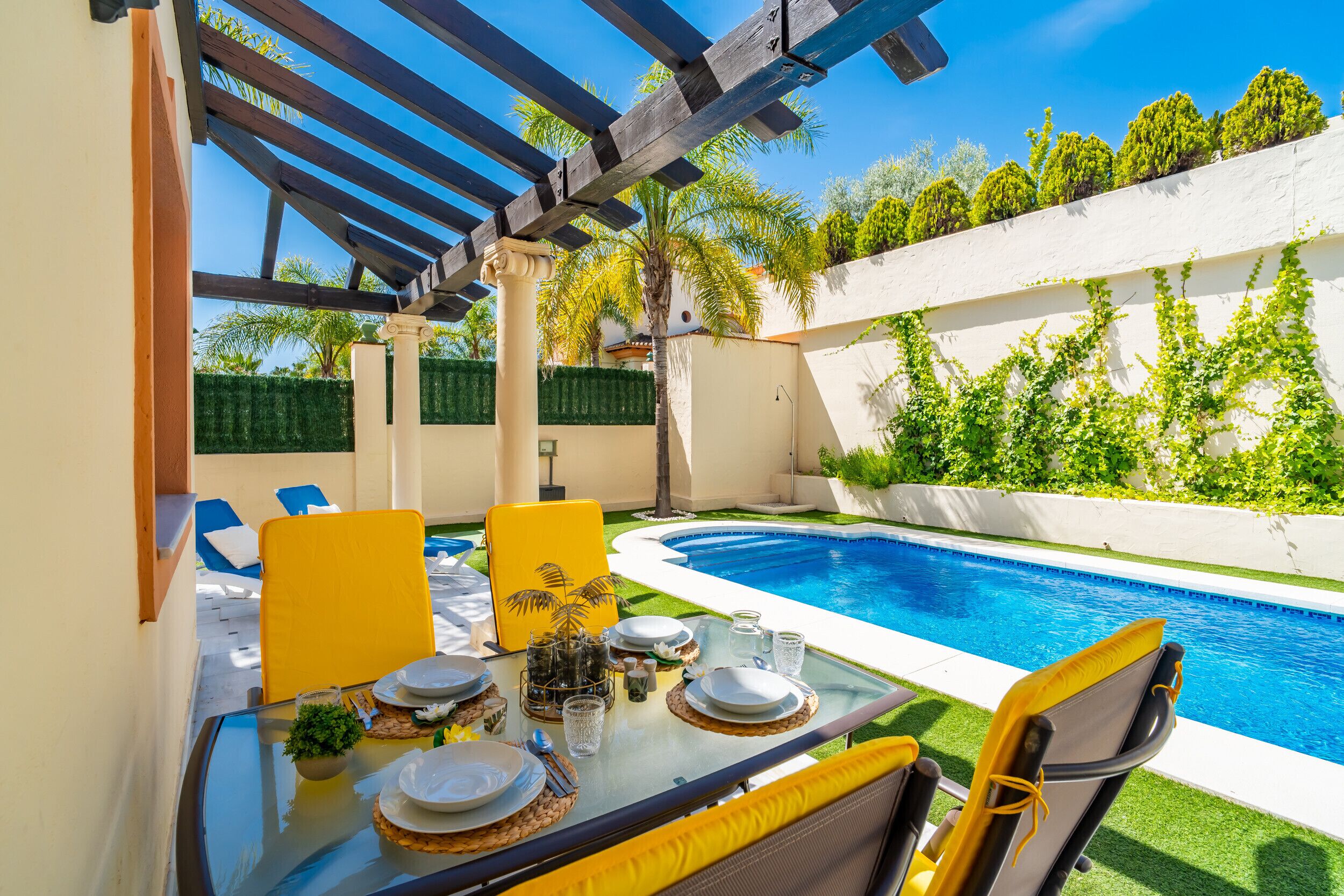 A poolside dining area with a table set for two, a pergola, and a view of the pool.