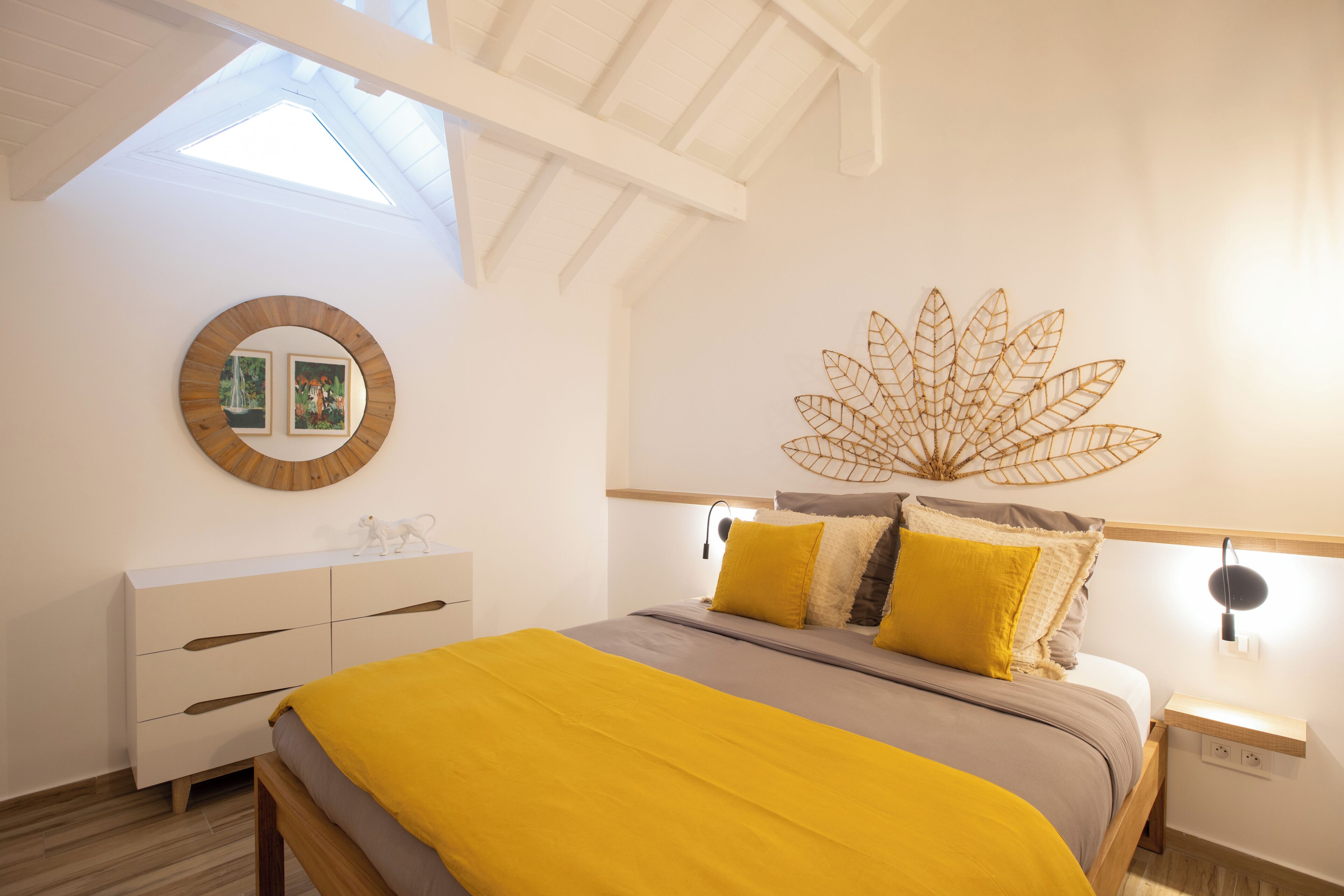 Un dormitorio con tragaluz, una cama con manta amarilla, un tocador blanco y una hoja decorativa en la pared.