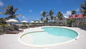 A pool area with lounge chairs and umbrellas, surrounded by palm trees and a building with a red roof.