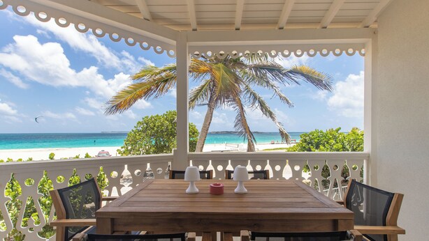 A wooden table with four chairs, two white lamps, and a red candle on a balcony with a white railing and decorative elements. A palm tree and a beach with clear blue water are visible in the background.