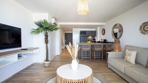 A modern living room with a flat-screen TV, a round coffee table, a sofa, a dining area with chairs, and a kitchen in the background.