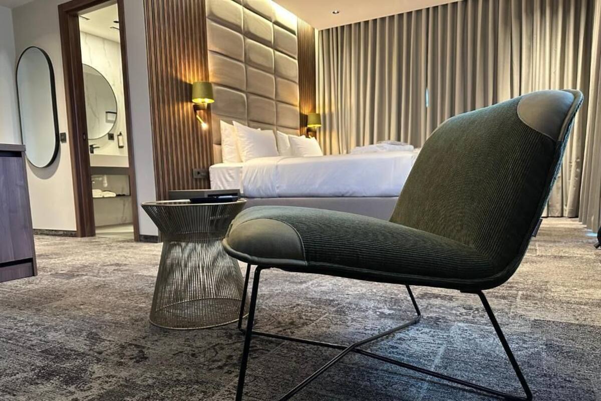 Ein modernes Hotelzimmer mit einem großen Bett, einem grünen Polsterstuhl, einem runden Spiegel und einem kleinen Nachttisch.