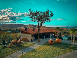 Un edificio rústico de piedra con tejado de baldosas, un gran árbol en primer plano y vistas de montañas distantes.